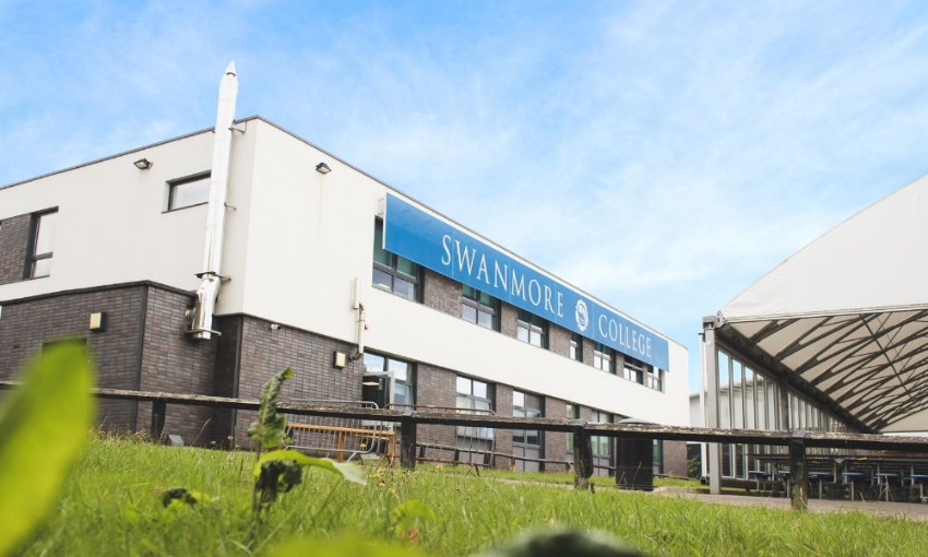 Low-angle view of the modern exterior of Swanmore College featuring a blue sign on a white facade, a dark brick base, and a grassy foreground.
